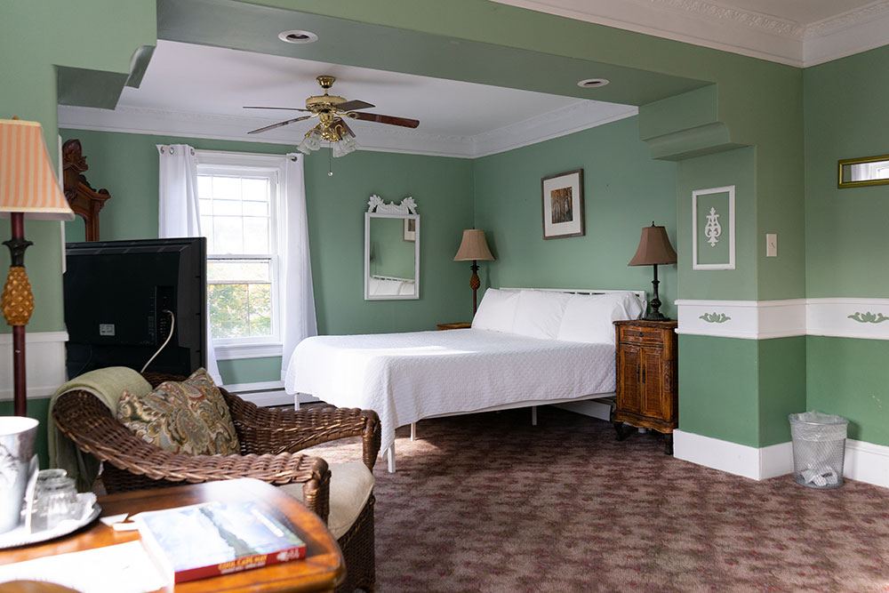 Commodore John Jacoby room with sage-green walls, ceiling fan and a cane chair